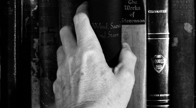 Black and white photo of books on a shelf with someone's hand reaching for a book