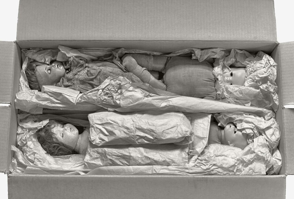 Black and white photo of cardboard box with dolls packed in it