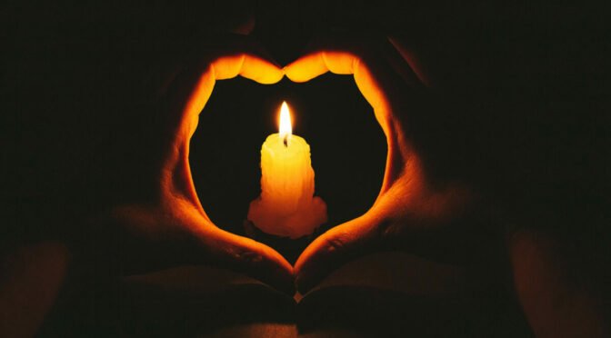 Photo of hands in shape of heart around candle in dark