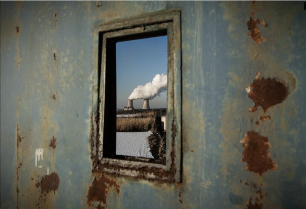 Photo of old, rusty wall with a window, smoke stacks billowing seen through the window