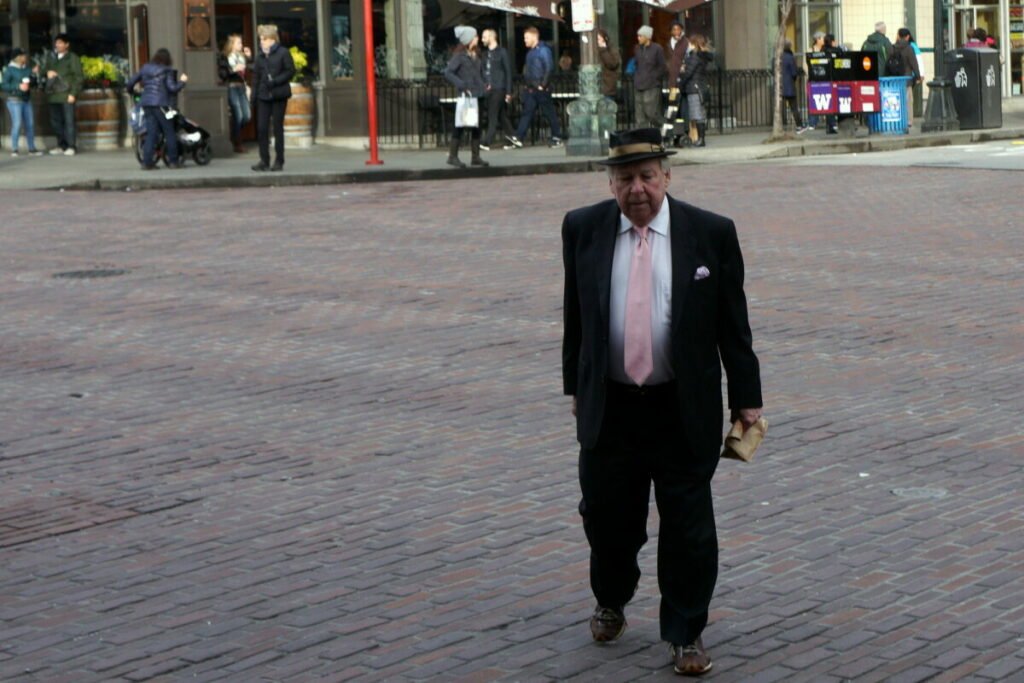 Elder man in fedora and pink tie crossing a brick street