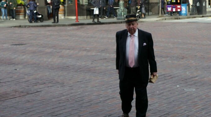 Elder man in fedora and pink tie crossing a brick street