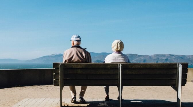 Photo from behind of older couple sitting on bench in front of mountains
