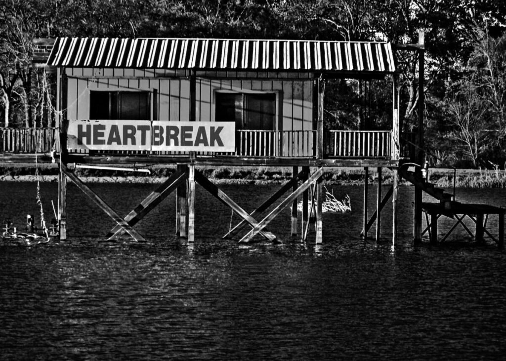 Black and white photo of small house on water with a sign outside that says "Heartbreak"