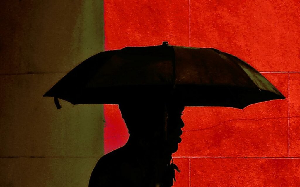 Photo of silhouette of man under umbrella, with a red wall meeting a beige wall behind him