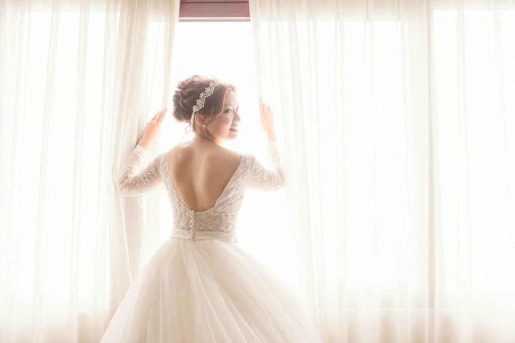Photo of woman in wedding dress looking over her shoulder, opening curtains to bright day