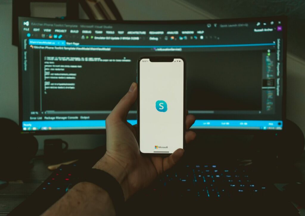 Photo of a hand holding a phone up in front of a computer screen with Skype icon open
