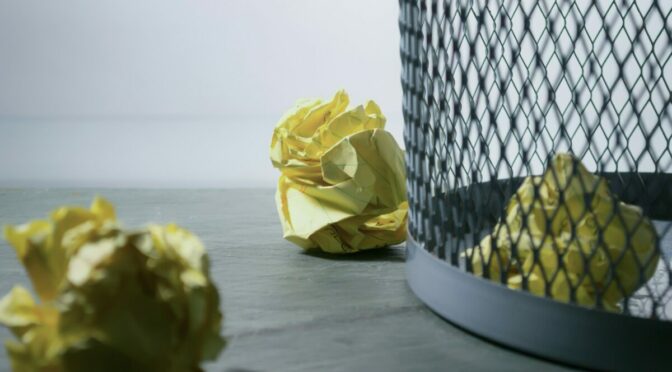 three crumpled pages of yellow paper and mesh trash can