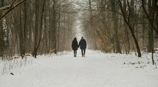 Photo of couple walking in snow under bare trees